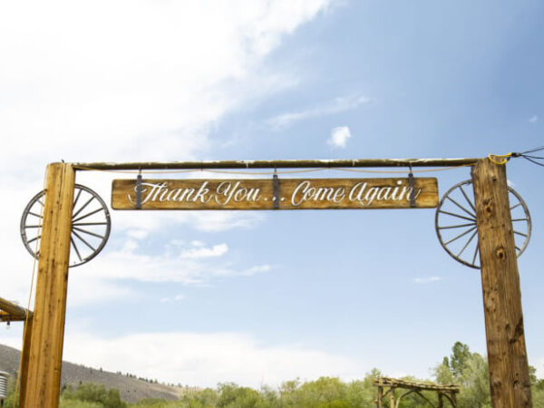 Wooden archway with a sign reading "Thank You... Come Again" under a clear sky, bordered by two wagon wheels, set against the picturesque backdrop of Bishop, California in the Eastern Sierra. visit bishop