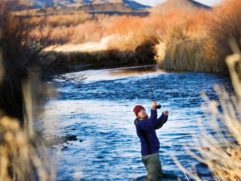 A person in outdoor clothing and a red beanie stands in a river near Bishop, California, casting a fishing line. Surrounding them are tall grasses and shrubs, with mountains visible in the background under a clear blue sky. visit bishop