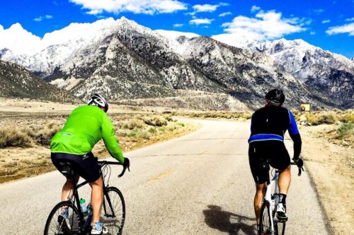 Two cyclists ride along an empty road towards snow-capped mountains under a bright blue sky with scattered clouds near Bishop, California. One cyclist wears a green jacket and the other sports a black and blue outfit. The scene is surrounded by rocky terrain and dry vegetation. visit bishop