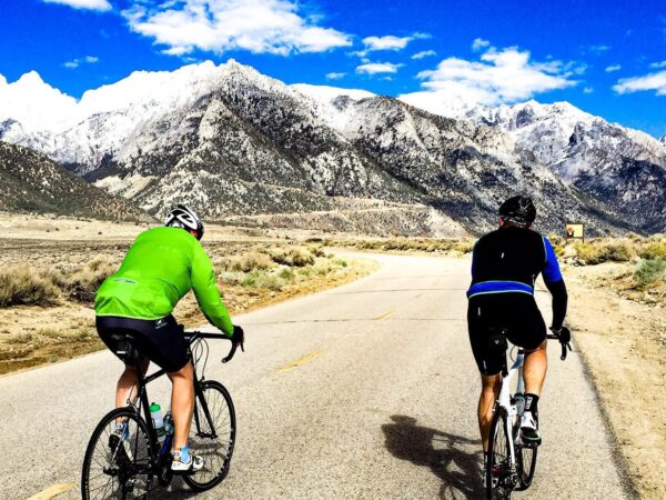 Two cyclists ride along an empty road towards snow-capped mountains under a bright blue sky with scattered clouds near Bishop, California. One cyclist wears a green jacket and the other sports a black and blue outfit. The scene is surrounded by rocky terrain and dry vegetation. visit bishop