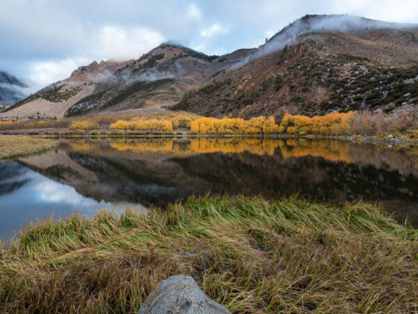 A serene mountain lake near Bishop, California reflects the rugged, tree-lined peaks and a line of yellow-leaved trees. The grassy foreground leads to calm waters, with scattered rocks and mist hovering near the mountaintops under a partly cloudy sky. visit bishop