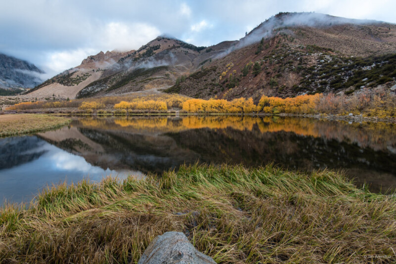 A serene mountain lake near Bishop, California reflects the rugged, tree-lined peaks and a line of yellow-leaved trees. The grassy foreground leads to calm waters, with scattered rocks and mist hovering near the mountaintops under a partly cloudy sky. visit bishop