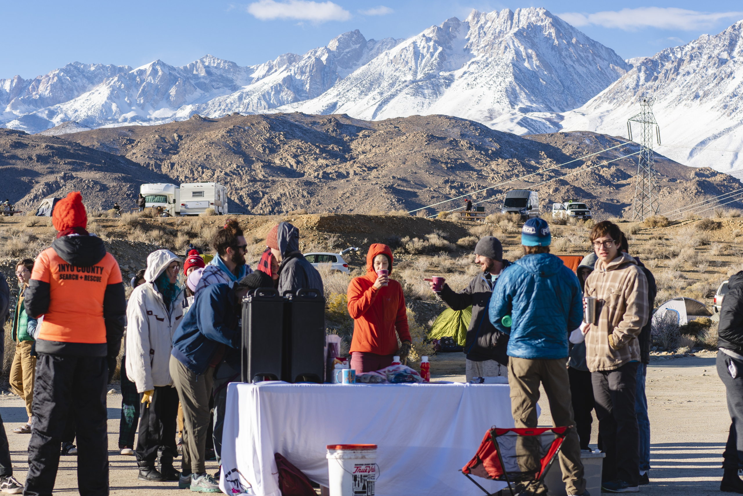 A group of people gathered around a table in the mountainous, snowy landscape of Bishop, California, with tents and RVs dotting the background of the stunning Eastern Sierra. visit bishop