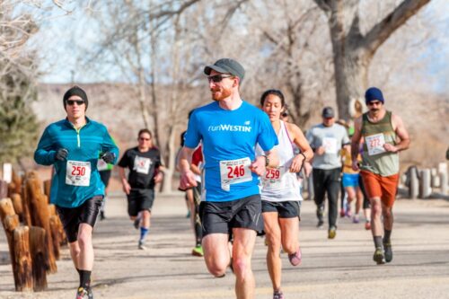 A group of runners jogs on a park trail during a race on a clear day in Bishop, California, with the stunning Eastern Sierra as their backdrop, all proudly wearing bib numbers. visit bishop