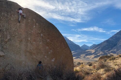 A person climbs a large boulder in the mountainous desert landscape of Bishop, California under a partly cloudy blue sky. visit bishop