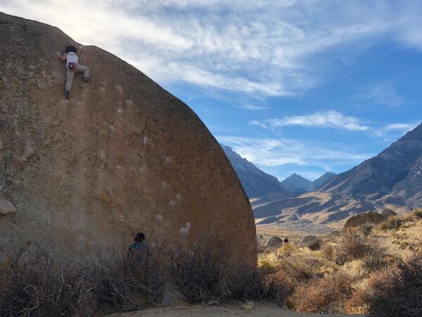 A person climbs a large boulder in the mountainous desert landscape of Bishop, California under a partly cloudy blue sky. visit bishop