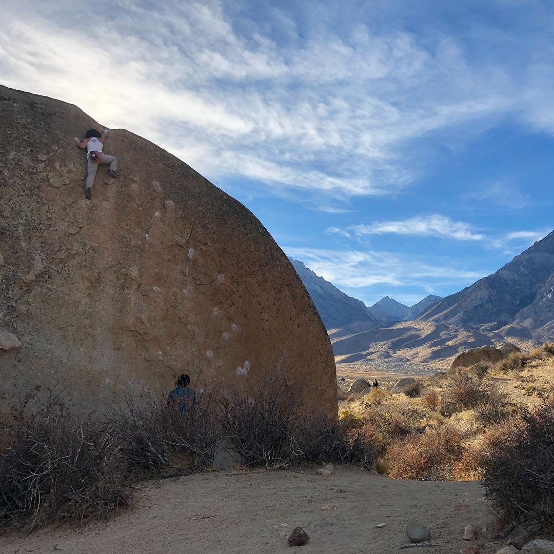 A person climbs a large boulder in the mountainous desert landscape of Bishop, California under a partly cloudy blue sky. visit bishop