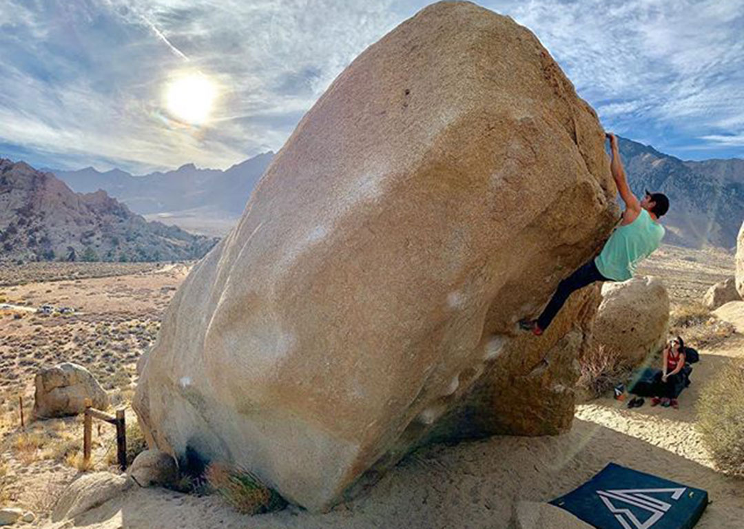 A person bouldering on a large rock with a rugged landscape, surrounded by mountains and the bright sun in the background, captures the adventurous spirit of Bishop, California in the Eastern Sierra. visit bishop