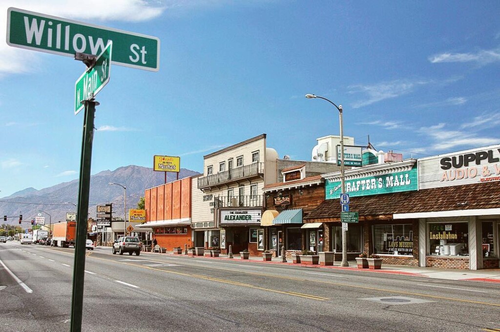 A small-town street in Bishop, California, features various shops and a "Willow St" sign in the foreground, with the majestic Eastern Sierra mountains visible in the background. visit bishop