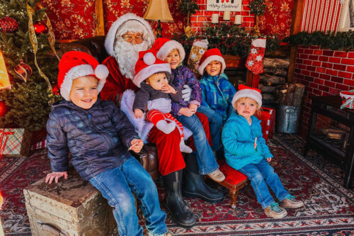 Five children in Santa hats sit with Santa in a festive Christmas setting in Bishop, California. Two trees and red decorations are visible, capturing the holiday spirit of the Eastern Sierra. visit bishop