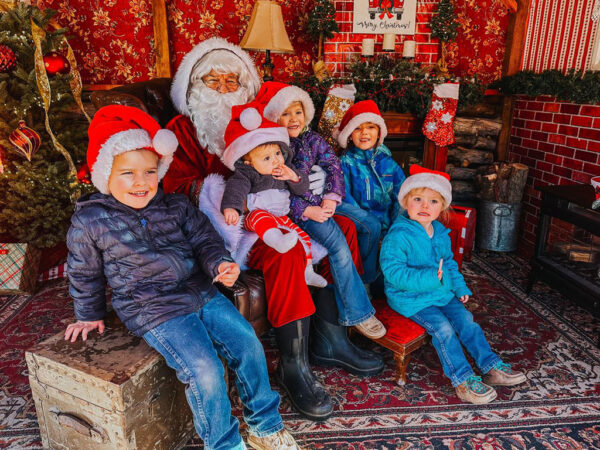 Five children in Santa hats sit with Santa in a festive Christmas setting in Bishop, California. Two trees and red decorations are visible, capturing the holiday spirit of the Eastern Sierra. visit bishop