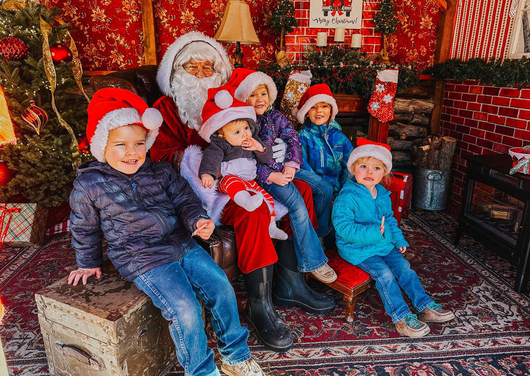 Five children in Santa hats sit with Santa in a festive Christmas setting in Bishop, California. Two trees and red decorations are visible, capturing the holiday spirit of the Eastern Sierra. visit bishop