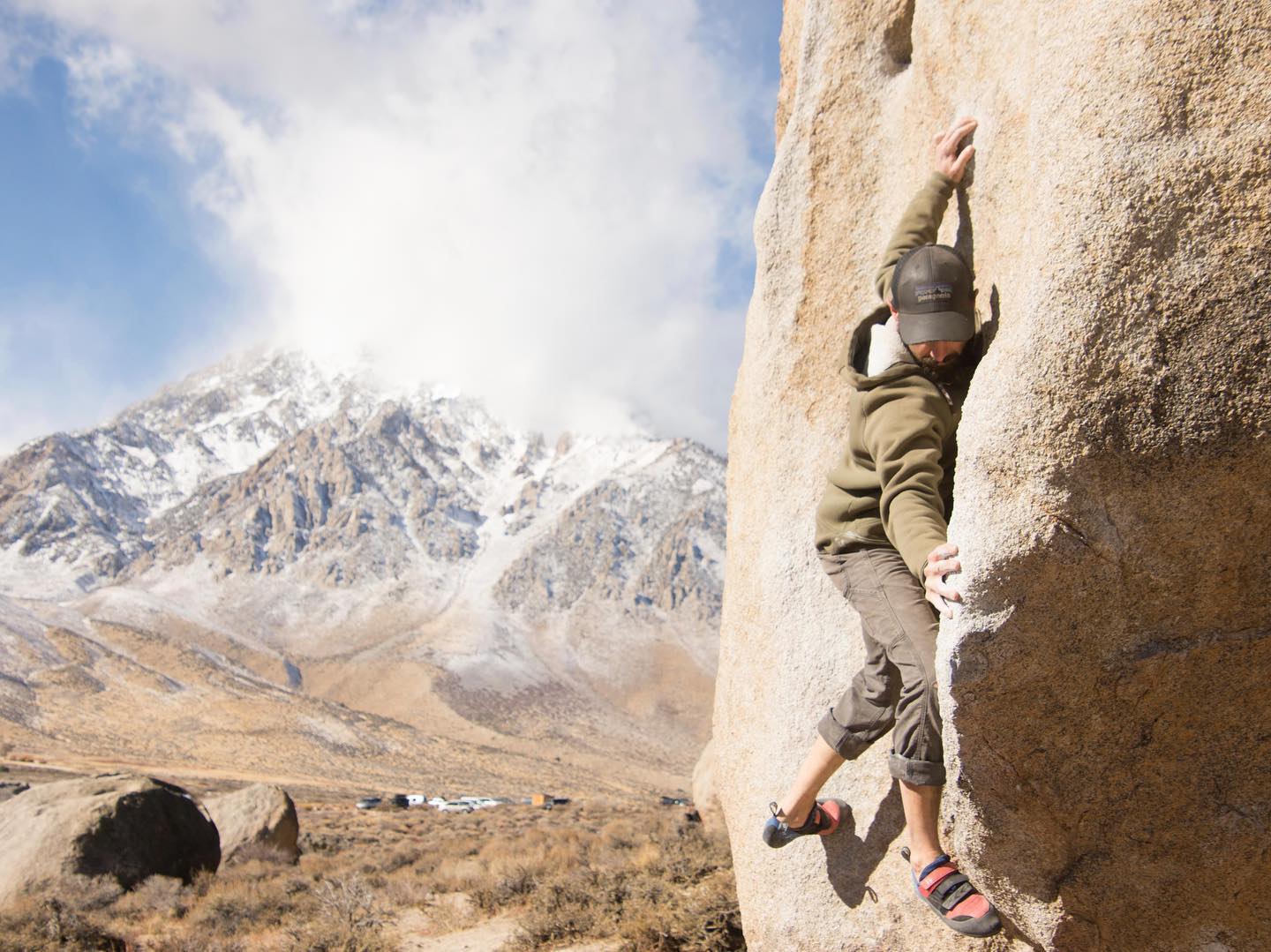 A person climbing a boulder in a mountainous landscape, with the snow-capped peaks of the Eastern Sierra in the background and the sky partly cloudy. visit bishop