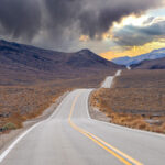 A winding road through the vast, barren landscape of Bishop, California, with dark clouds overhead and a glimpse of sunset on the horizon in the Eastern Sierra. visit bishop