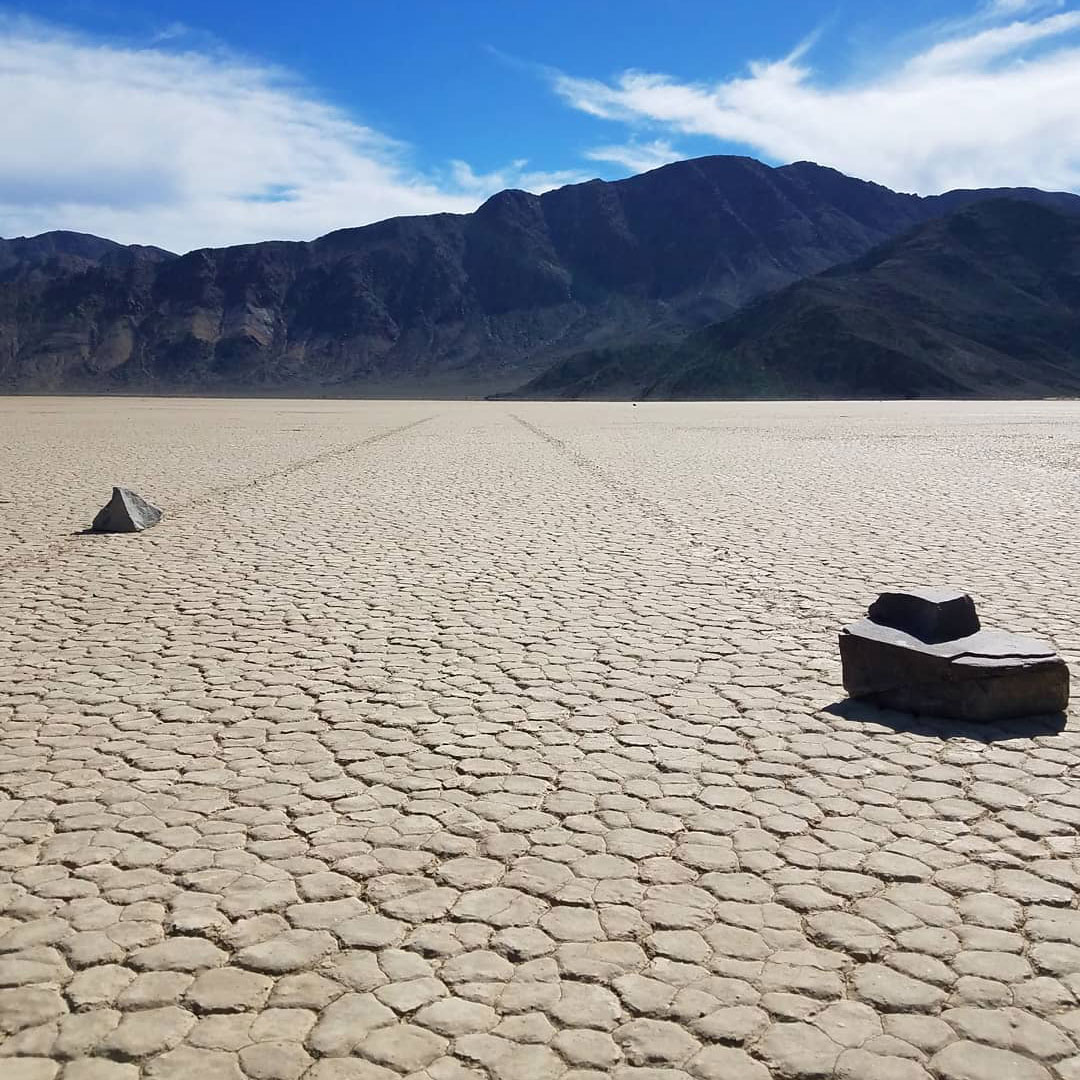 Large rocks on cracked desert ground with mountains in the background and a bright blue sky with scattered clouds, capturing the rugged beauty of Bishop, California, nestled in the heart of the Eastern Sierra. visit bishop