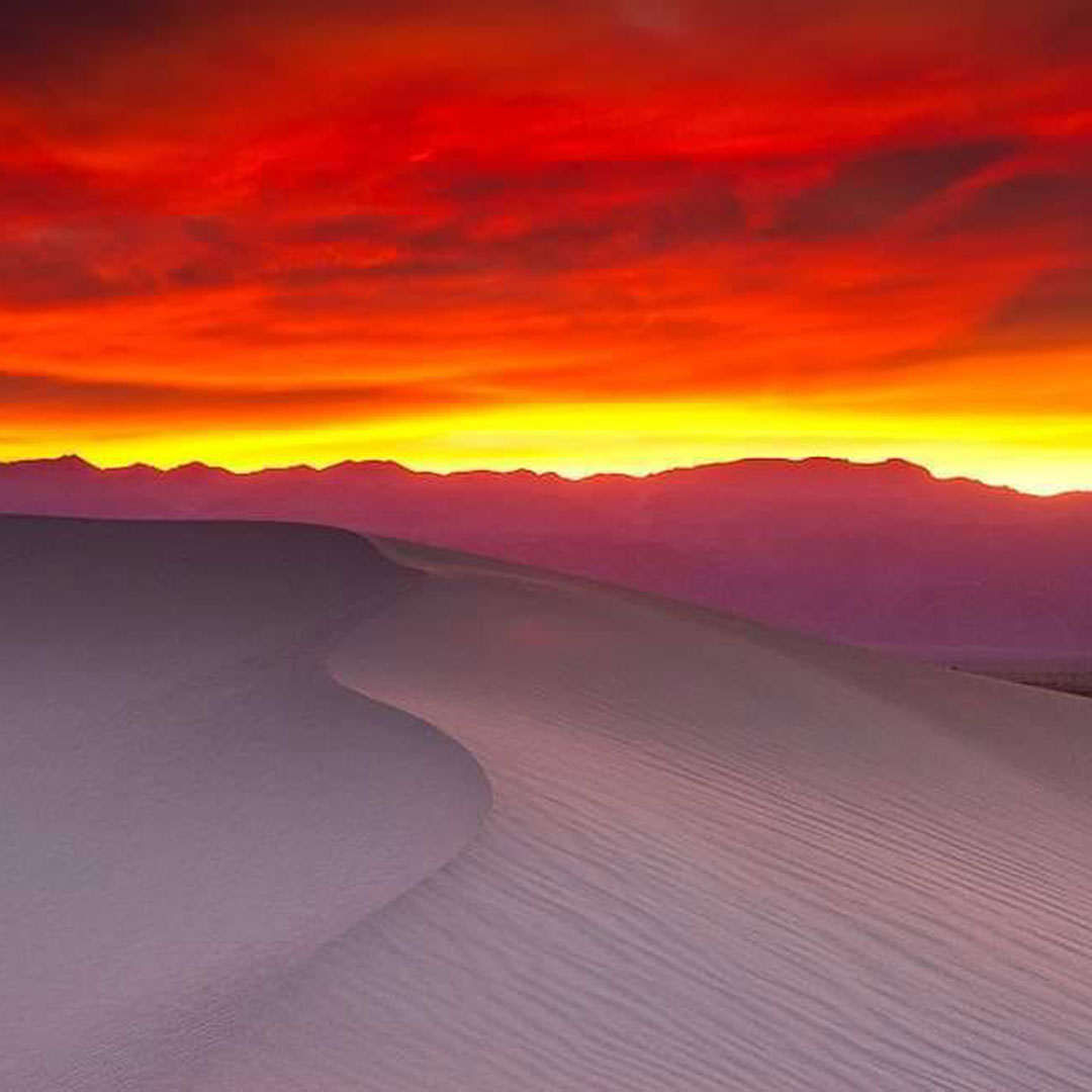 A tranquil desert scene at sunset showcases vibrant orange and red skies above the smooth sand dunes near Bishop, California, with the majestic Eastern Sierra in the distance. visit bishop