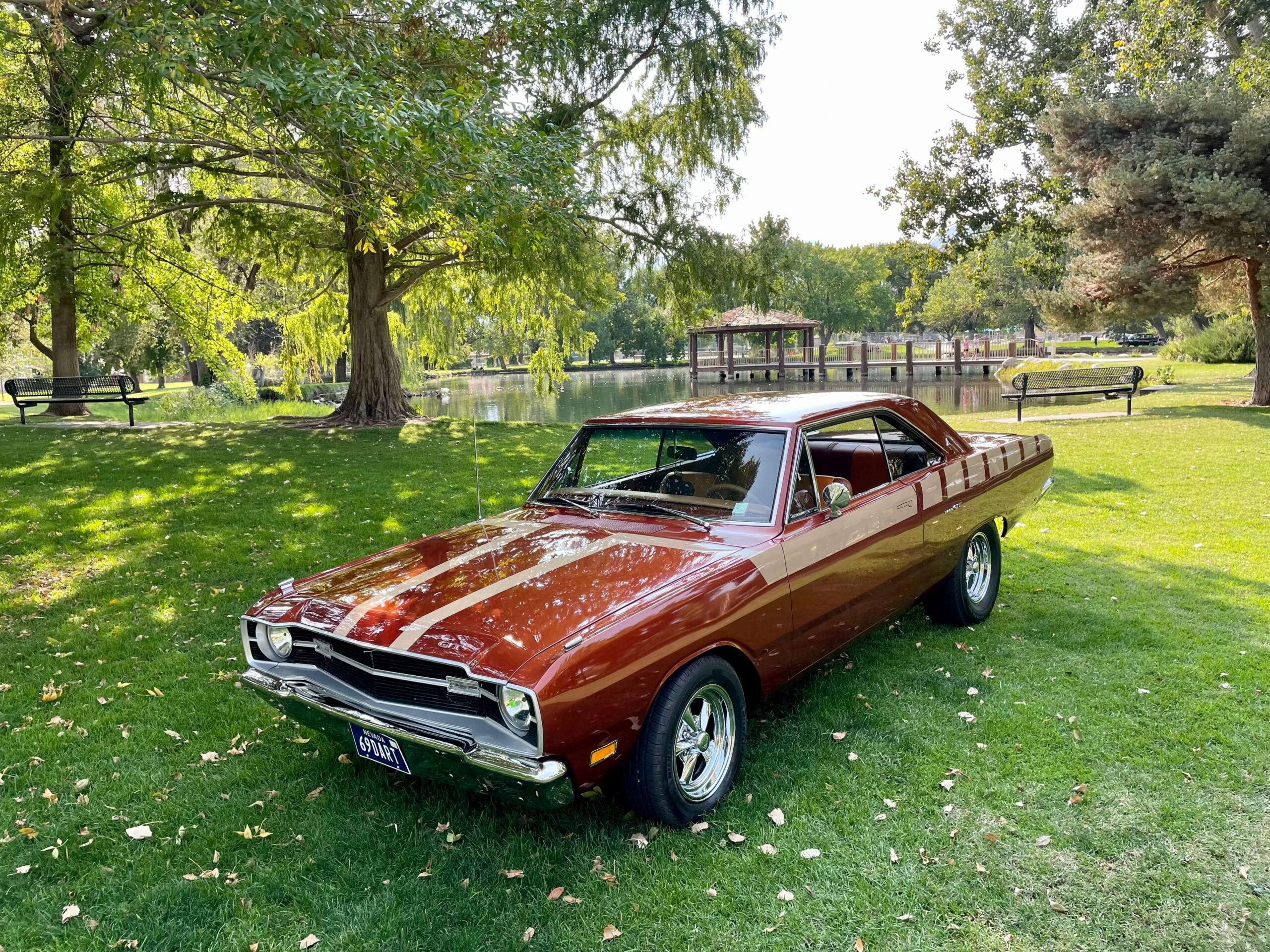 A vintage red car with white stripes parked on a green lawn near a pond and large trees, reminiscent of the serene beauty found in Bishop, California. visit bishop