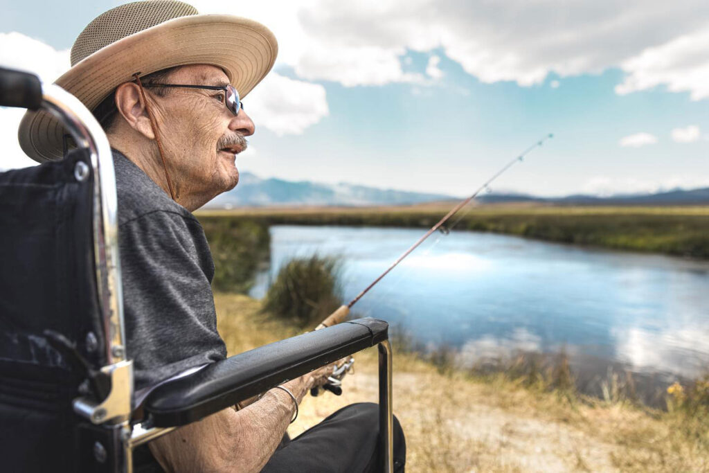 An elderly man in a wheelchair, wearing a hat and glasses, fishes by a serene lake in Bishop, California under the cloudy sky of the Eastern Sierra. visit bishop