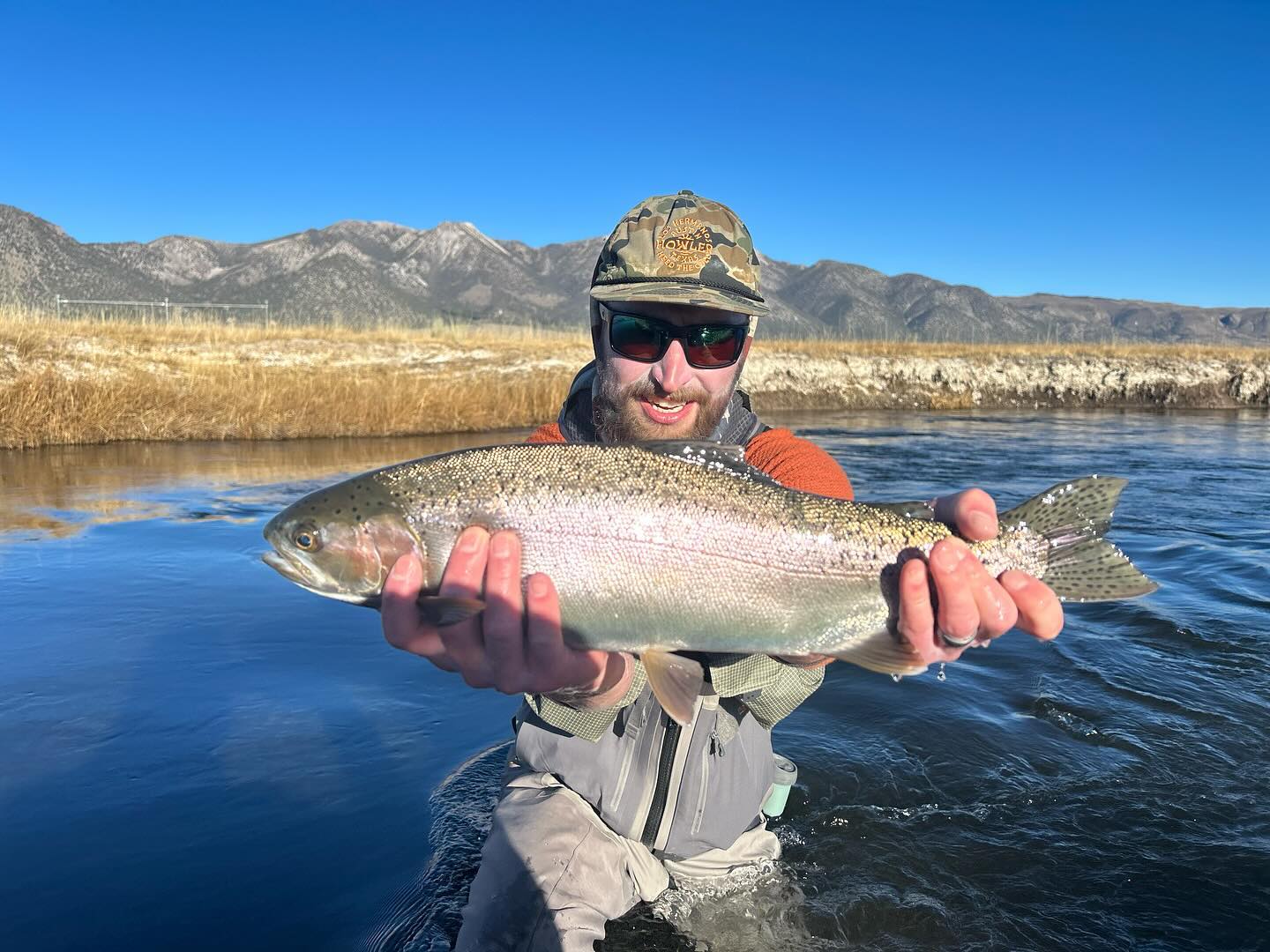 Man holding a large fish in a river with mountains in the background on a clear, sunny day near Bishop, California. visit bishop