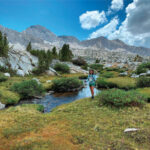 A person hiking near a stream in the Eastern Sierra of Bishop, California, navigates through a mix of greenery and rocky terrain under a cloudy sky. visit bishop