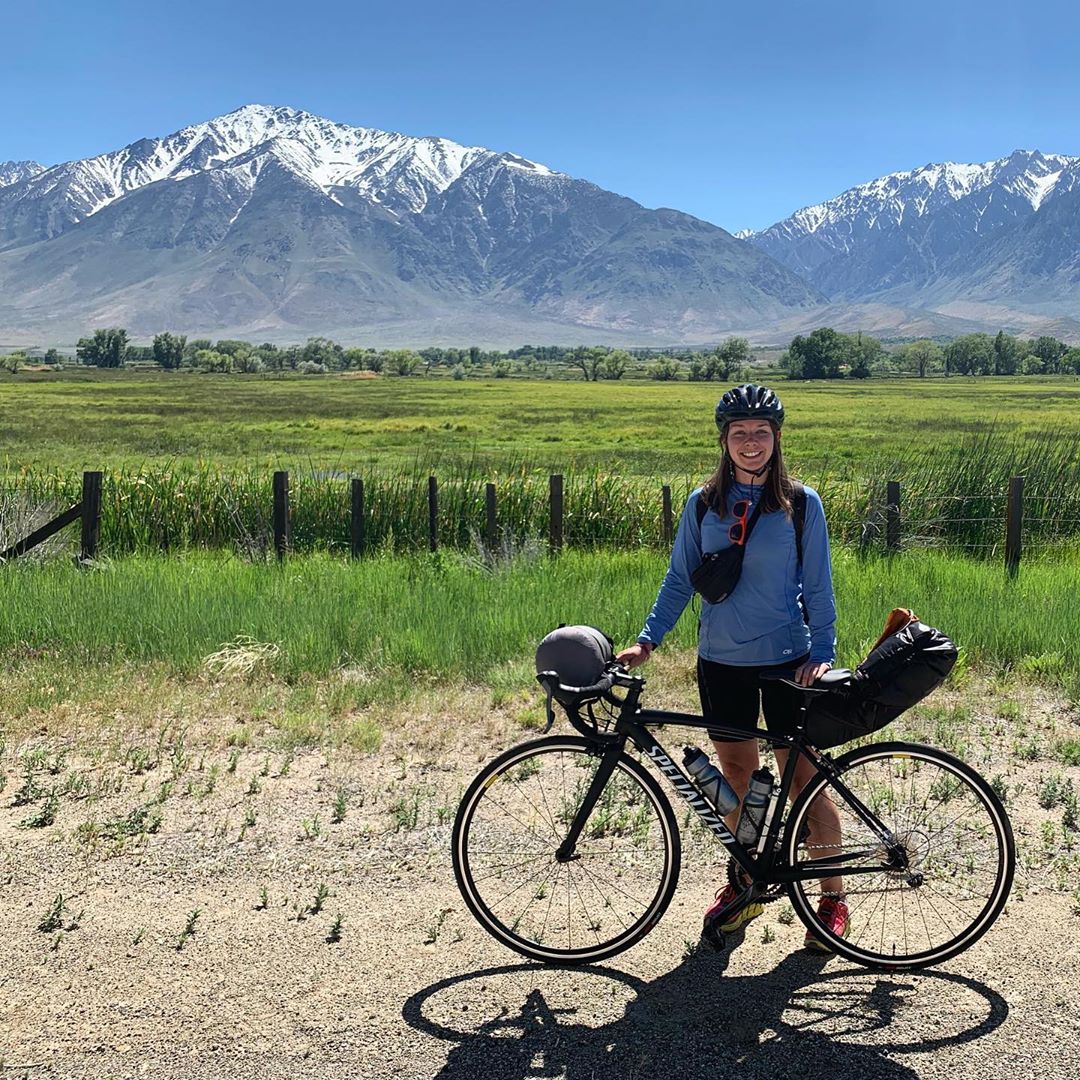 A person in biking gear stands with a bicycle in front of a scenic landscape with mountains and fields, capturing the stunning beauty of Bishop, California, nestled within the Eastern Sierra. visit bishop