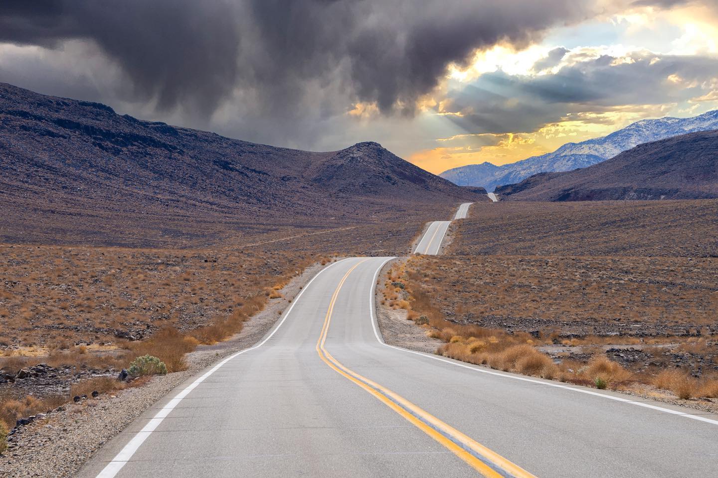 A winding road through a barren desert landscape with dramatic clouds and mountains in the background at sunset, capturing the rugged beauty of Bishop, California in the Eastern Sierra. visit bishop