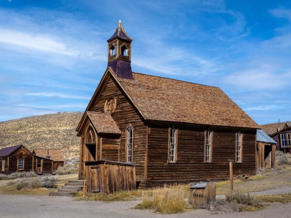 A wooden church in an old western-style ghost town near Bishop, California, with other wooden buildings in the background under a blue sky in the Eastern Sierra. visit bishop