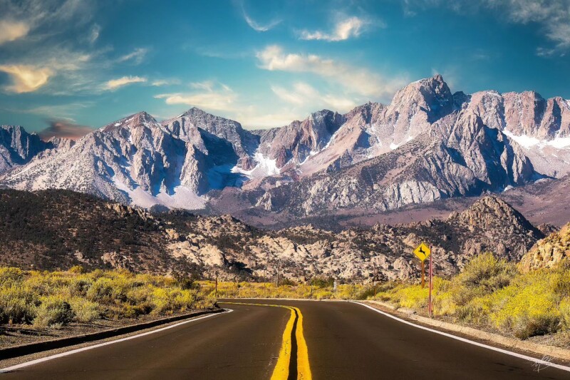 A winding road leading towards the rugged, snow-dusted mountains of the Eastern Sierra under a blue sky with wispy clouds near Bishop, California. visit bishop