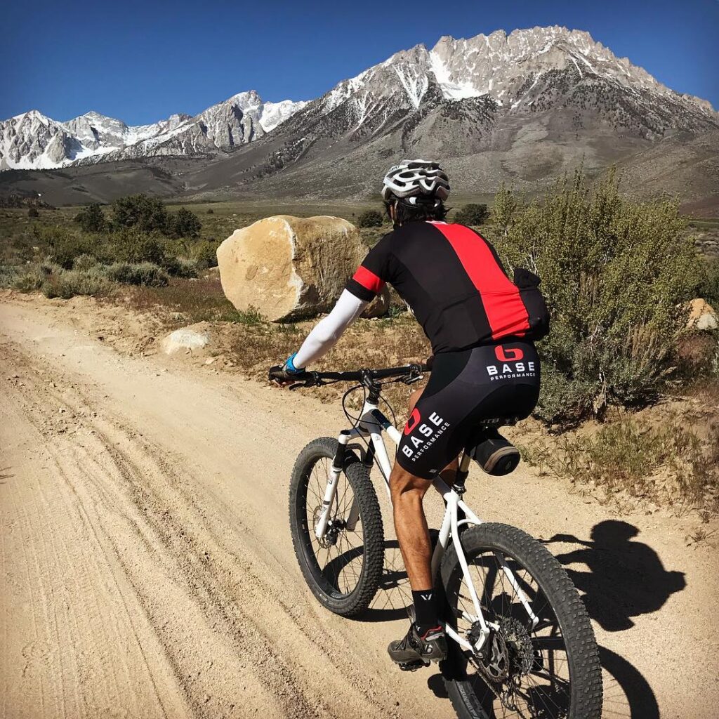 A cyclist in red and black gear rides on a dirt road with the stunning Eastern Sierra mountains and clear skies in the background, somewhere near Bishop, California. visit bishop