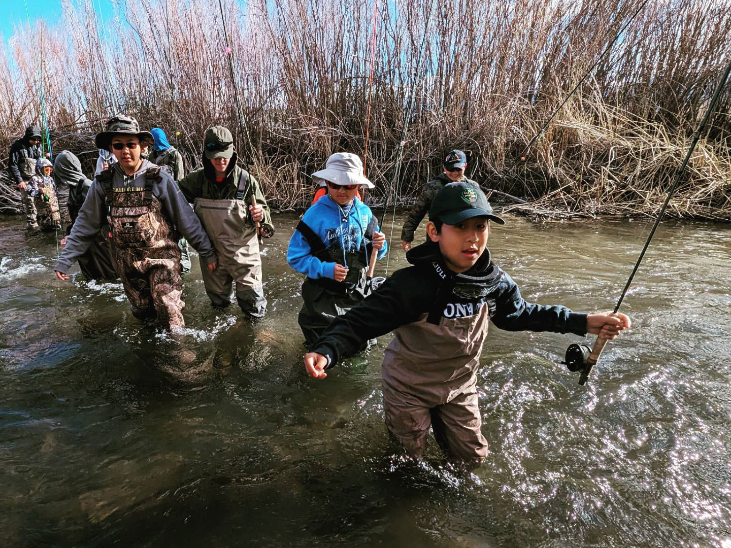 A group of kids wading through a river in Eastern Sierra, wearing waders and holding fishing rods, with tall reeds in the background. visit bishop