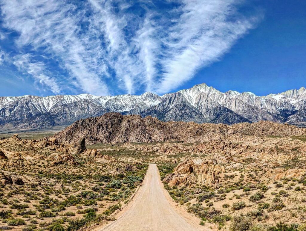 A dirt road leading towards snow-capped mountains in the Eastern Sierra under a sky streaked with clouds and blue hues. visit bishop