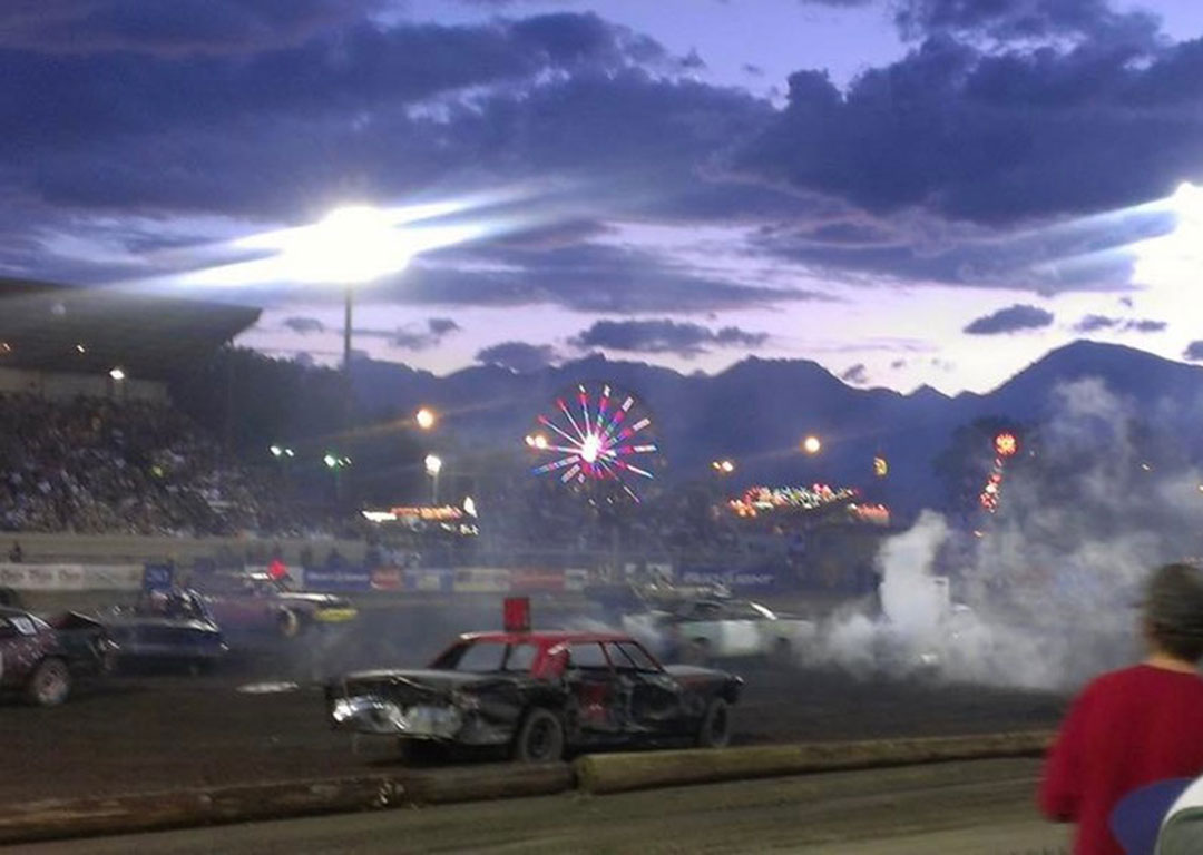 In Bishop, California, a nighttime demolition derby unfolds with cars crashing, smoke billowing, and a lit Ferris wheel in the background under the cloudy sky of the Eastern Sierra. visit bishop