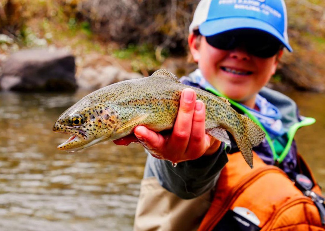 A person wearing a blue cap and sunglasses proudly holds a fish by the river in Bishop, California, against the stunning backdrop of the Eastern Sierra. visit bishop