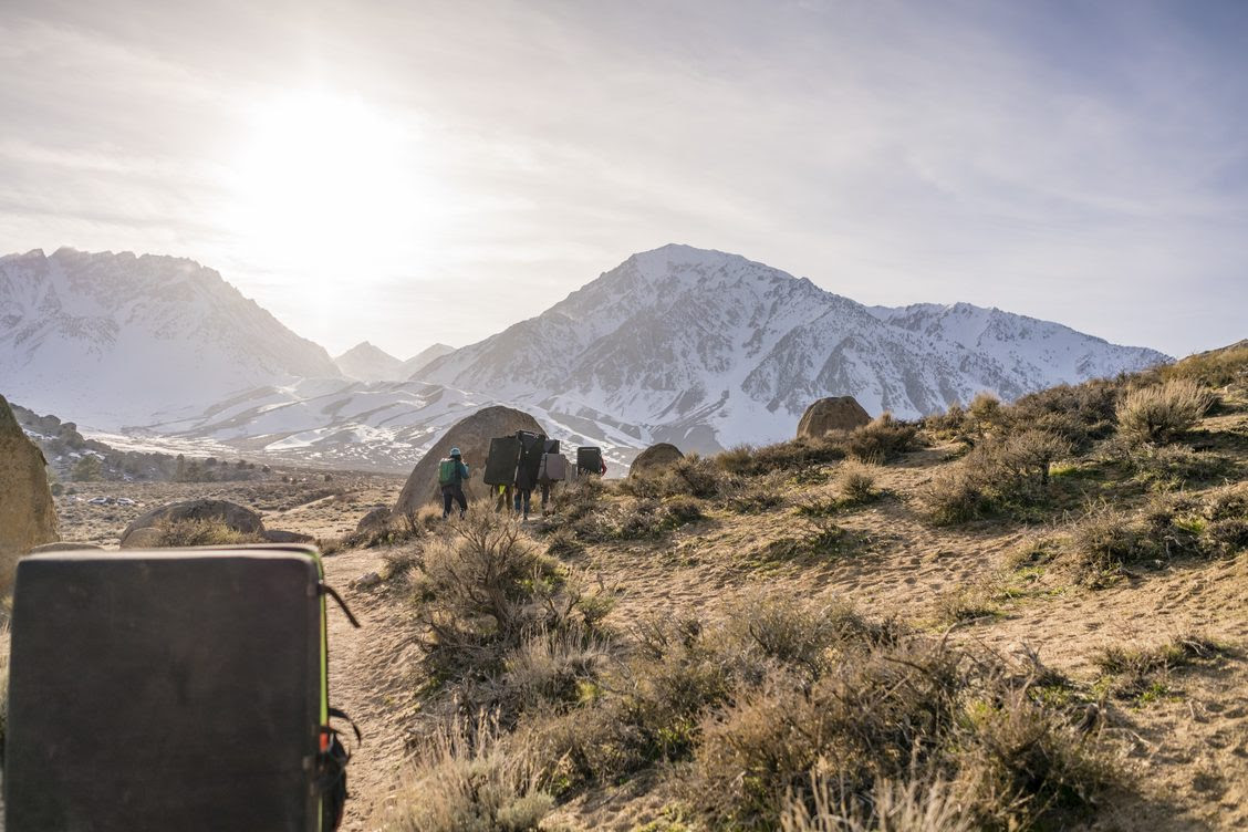 Three hikers with backpacks trek through a dry, rocky landscape near Bishop, California, with the snow-capped peaks of the Eastern Sierra in the background. visit bishop