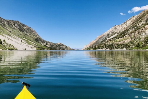 A yellow kayak glides on a calm lake surrounded by green mountains under a clear blue sky in Bishop, California's picturesque Eastern Sierra. visit bishop