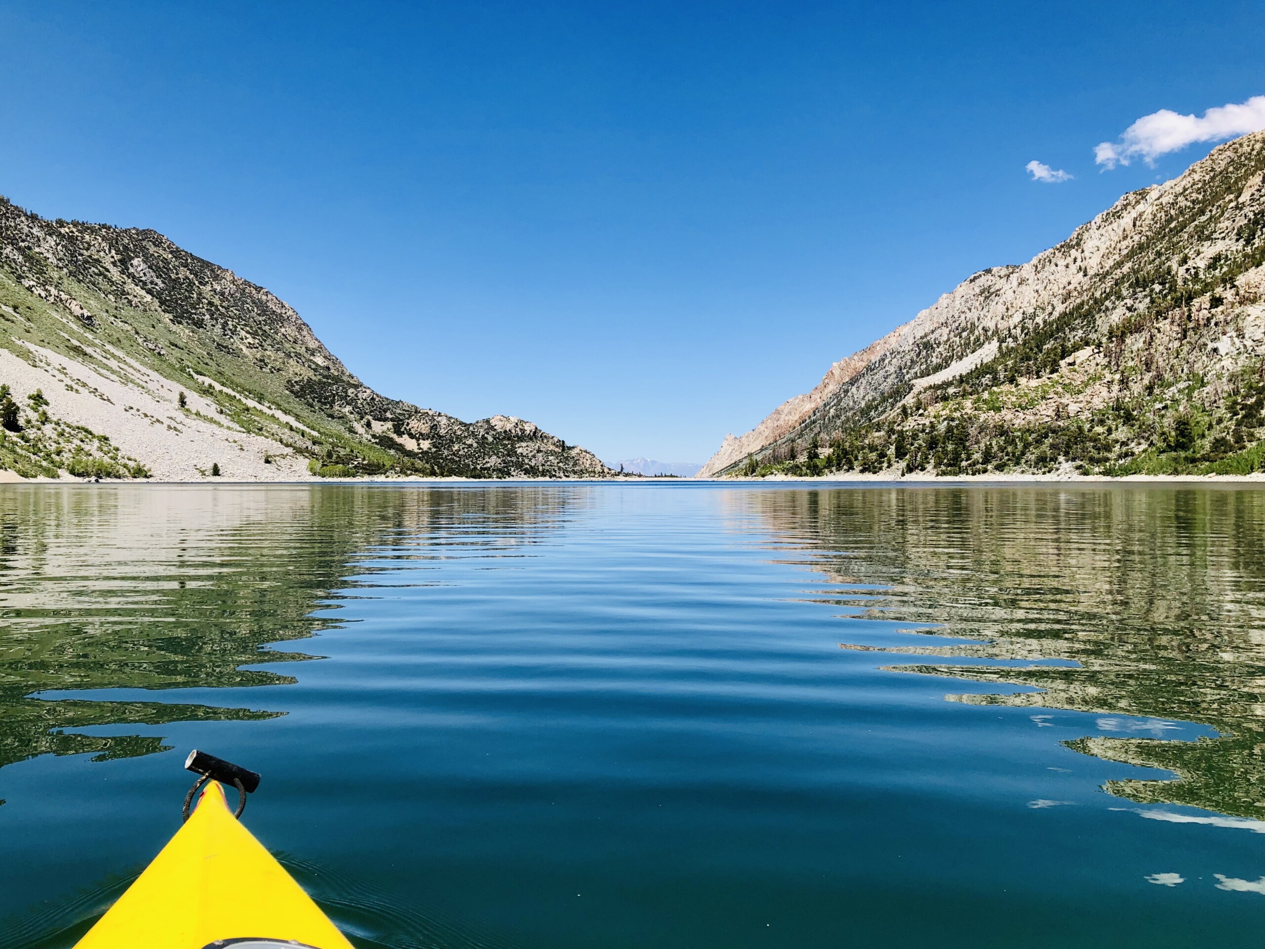 A yellow kayak glides on a calm lake surrounded by green mountains under a clear blue sky in Bishop, California's picturesque Eastern Sierra. visit bishop