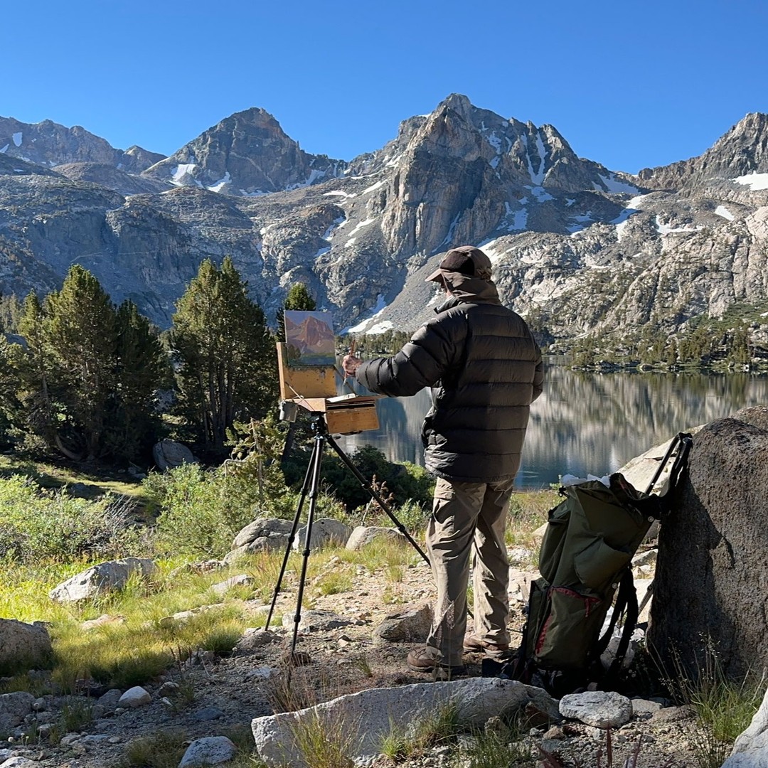 A person is painting a mountain landscape near a lake in the rugged terrain of the Eastern Sierra, standing next to their backpack and easel. visit bishop