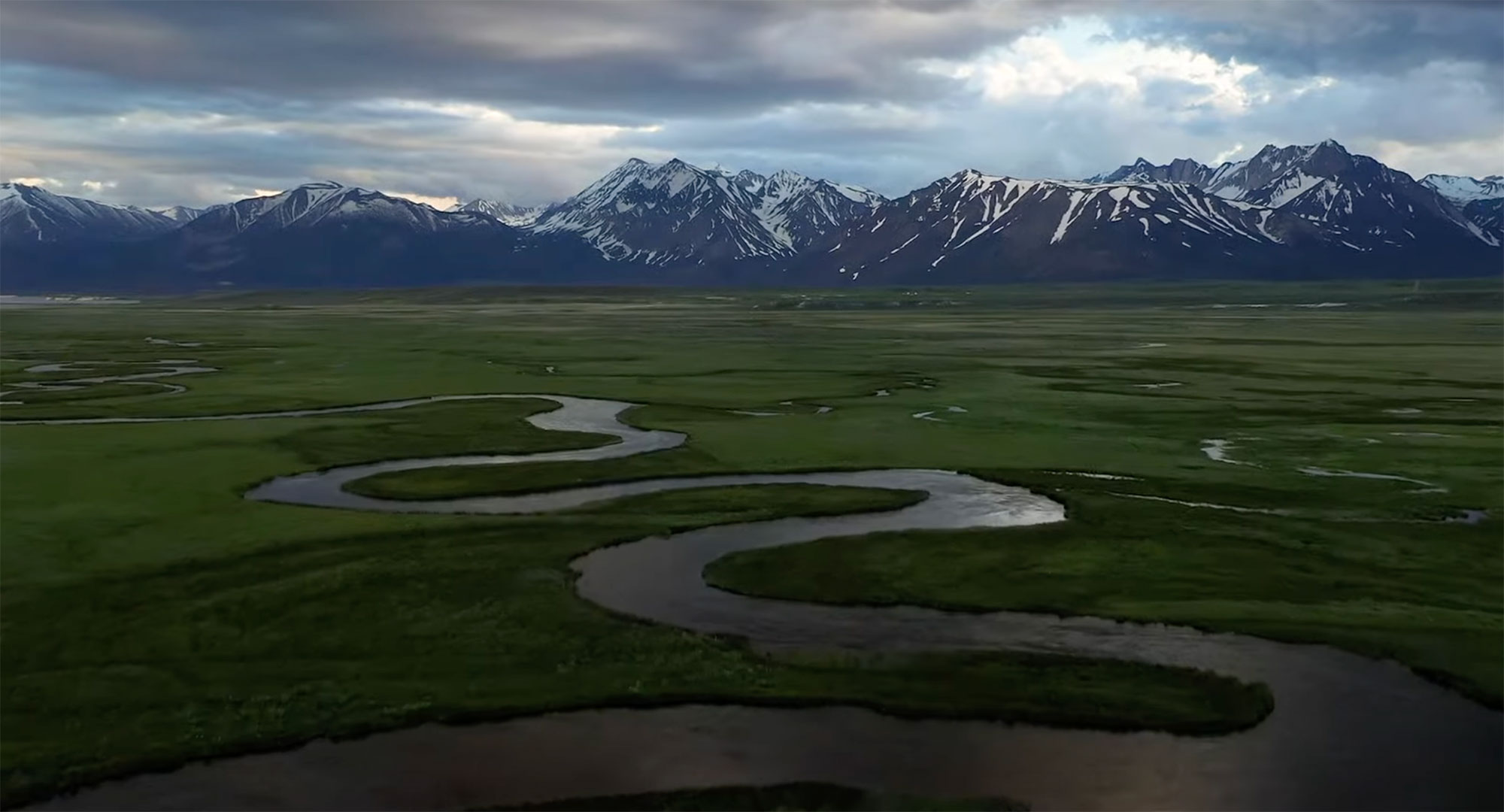 A winding river meanders through a green plain in Bishop, California, with the snow-capped peaks of the Eastern Sierra in the background under a cloudy sky. visit bishop