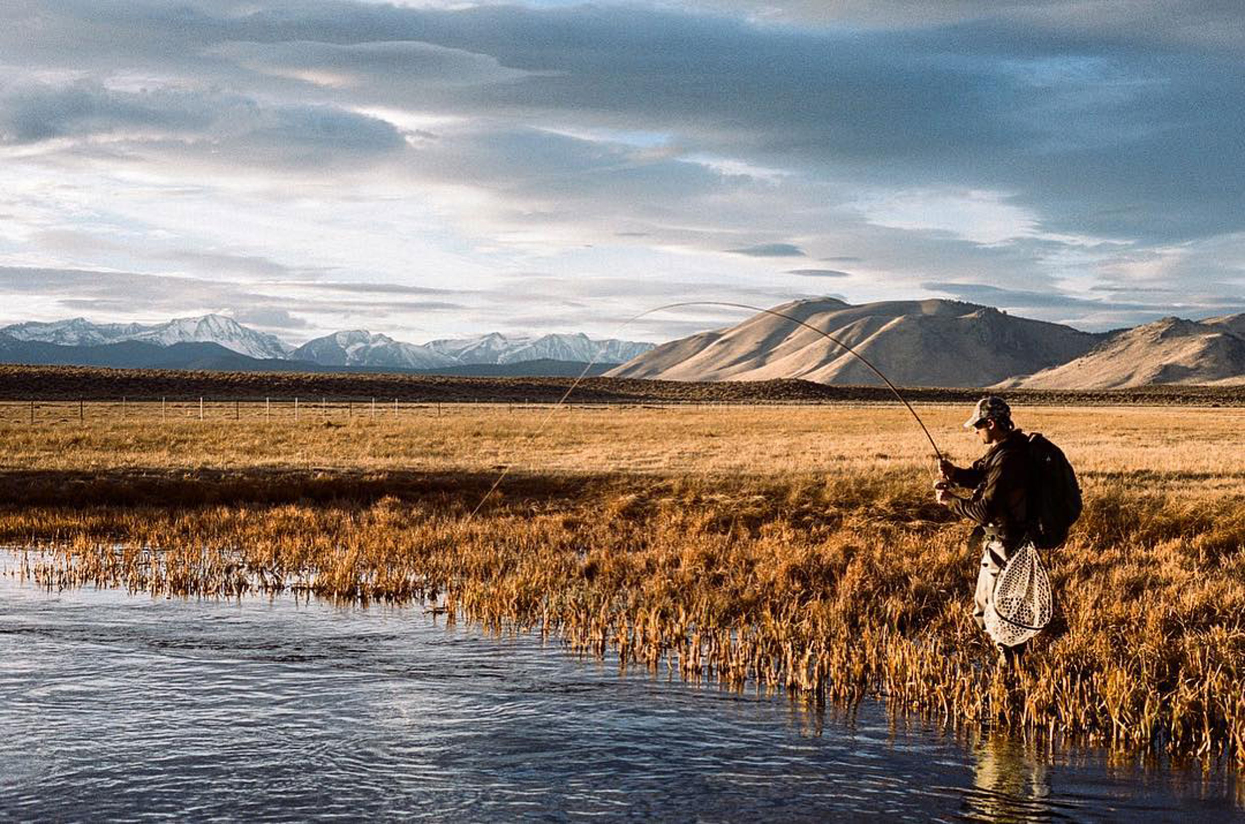 A person fly-fishing in a scenic field near a stream, with the majestic Eastern Sierra mountains and a partly cloudy sky in the background, just outside Bishop, California. visit bishop