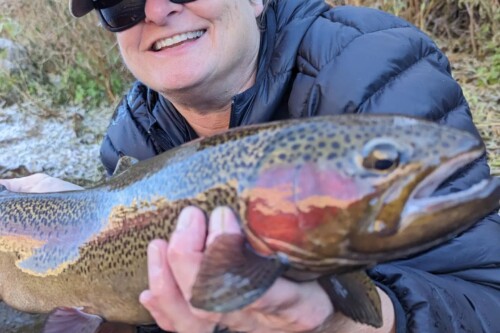 Person wearing sunglasses and a cap, smiling and holding a large trout near a stream in Bishop, California. visit bishop