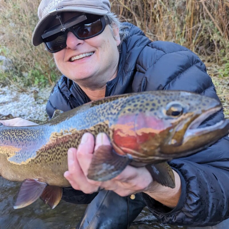 Person wearing sunglasses and a cap, smiling and holding a large trout near a stream in Bishop, California. visit bishop