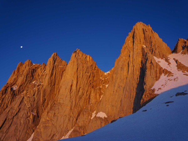 Jagged mountain peaks bathed in orange sunset light with a nearby moon against a dark blue sky and snowy foreground, capturing the breathtaking beauty of the Eastern Sierra near Bishop, California. visit bishop