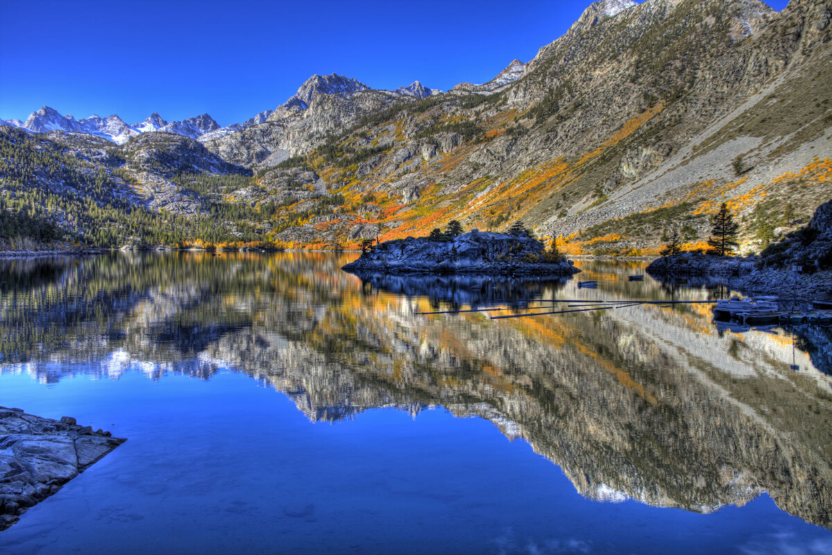 A calm mountain lake in Bishop, California, with clear reflections of the Eastern Sierra's rocky peaks and vibrant autumn foliage under a bright blue sky. visit bishop