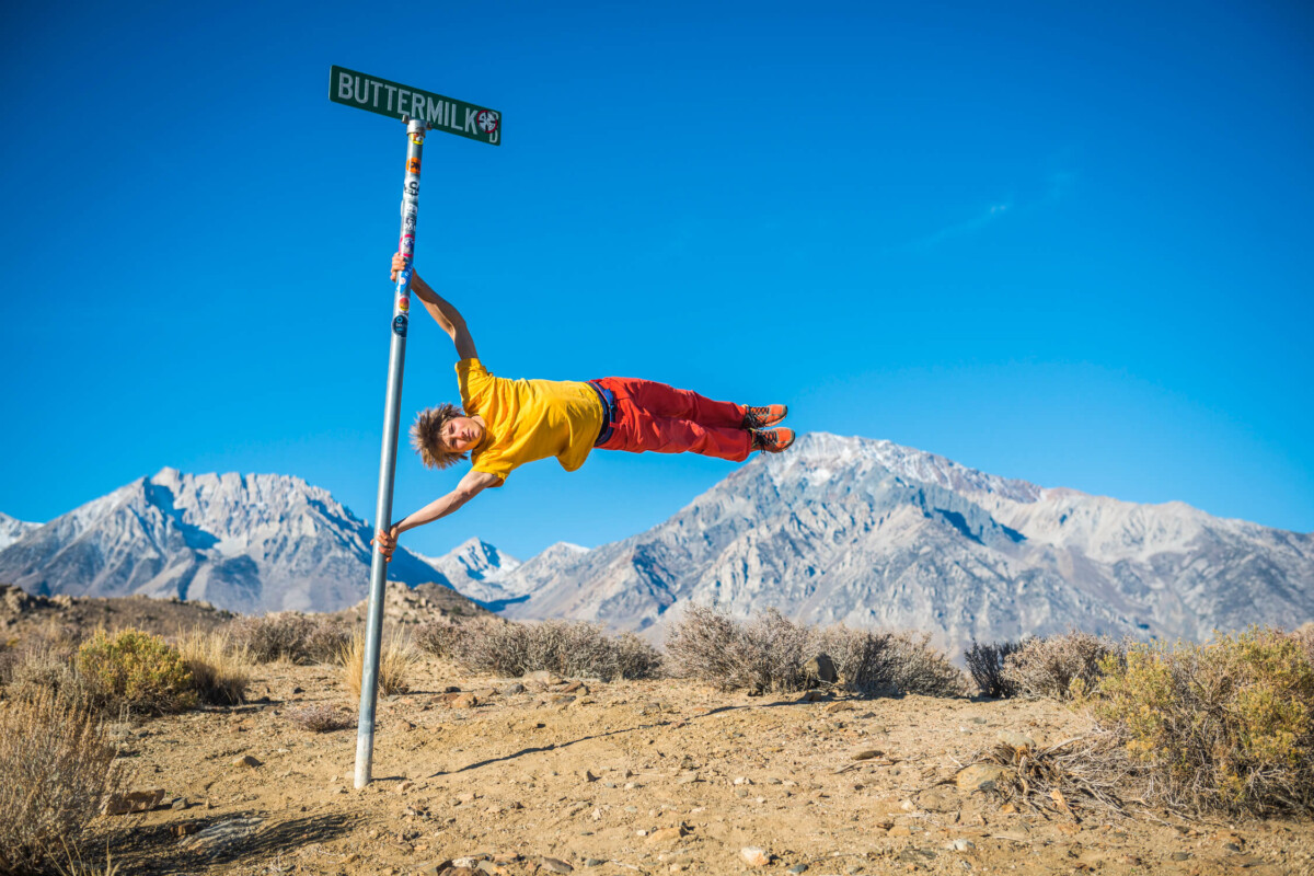 A person in a yellow shirt and red pants holds a Buttermilk signpost horizontally, with the majestic mountains of the Eastern Sierra near Bishop, California in the background. visit bishop