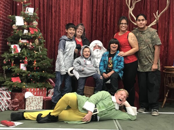 A family poses with Santa Claus and an elf in front of a decorated Christmas tree and a velvet curtain backdrop, capturing the holiday spirit in Bishop, California. visit bishop