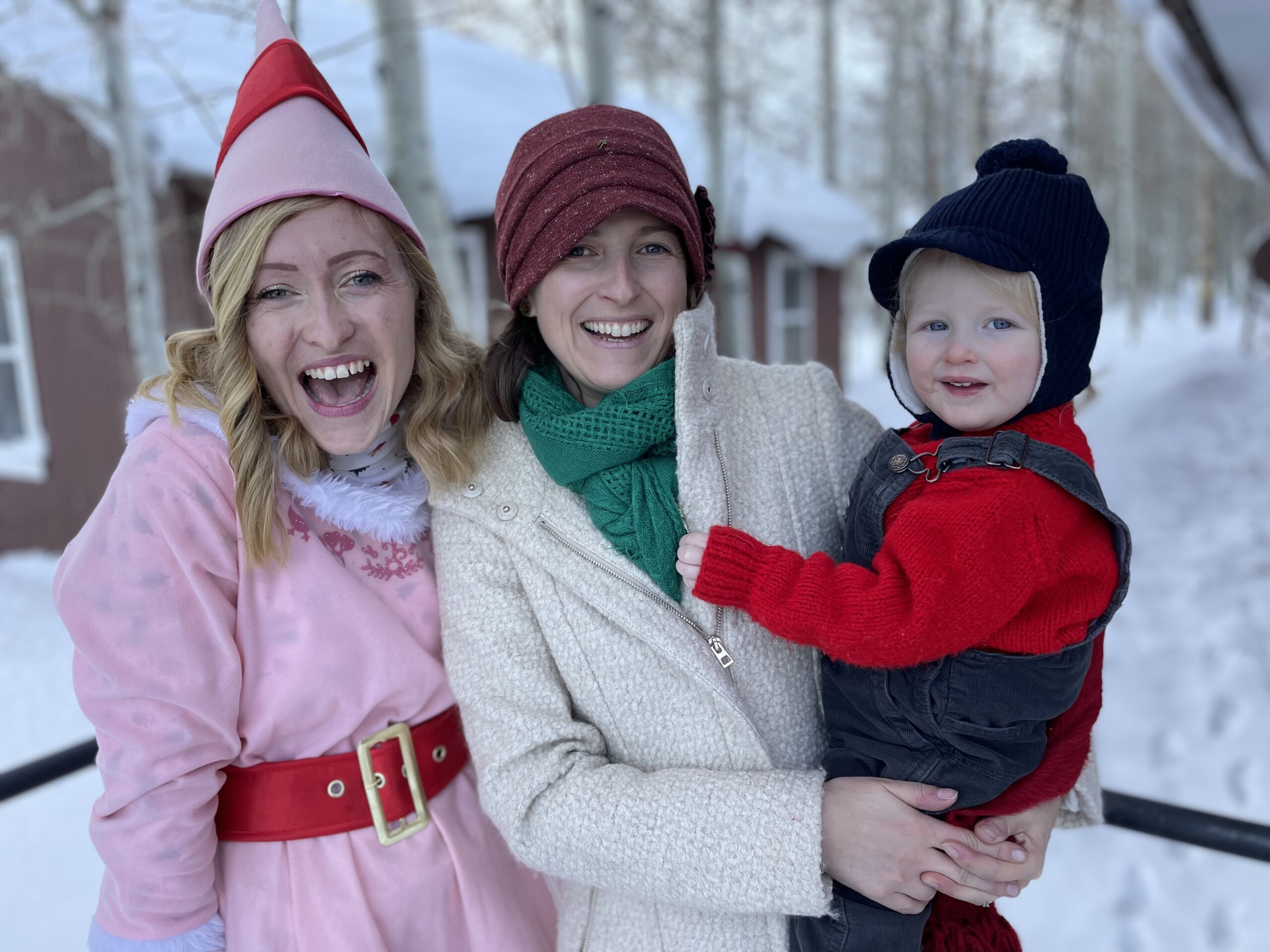 Three people in winter wear smile outdoors in a snowy setting in Bishop, California, including an elf costume, a white coat, and a red sweater. visit bishop