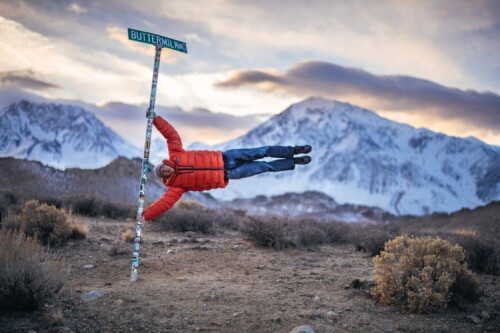 Person in a red jacket holds onto a signpost marked "Buttermilk Rd." while hovering horizontally, with the snowy mountains of the Eastern Sierra behind. visit bishop