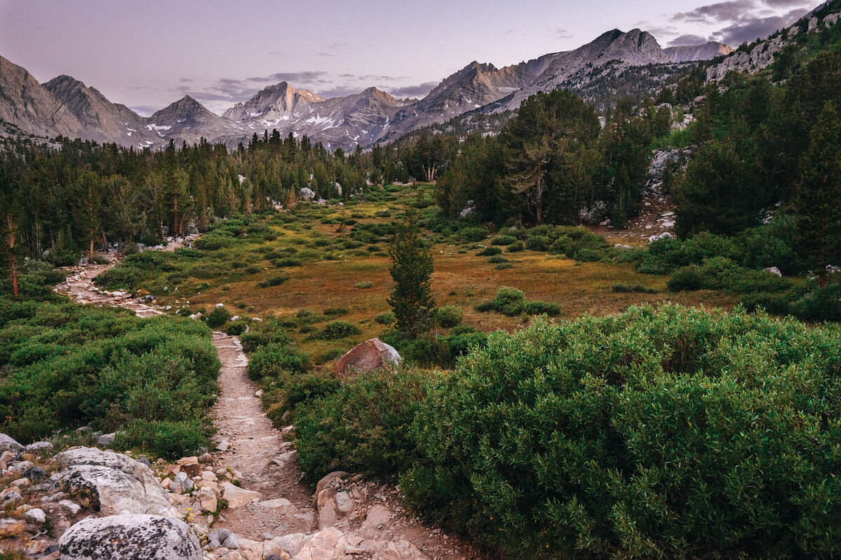A rocky trail leads into a green valley surrounded by the majestic mountains of Bishop, California, under a cloudy sky. Trees and bushes line the path, capturing the rugged beauty of the Eastern Sierra. visit bishop