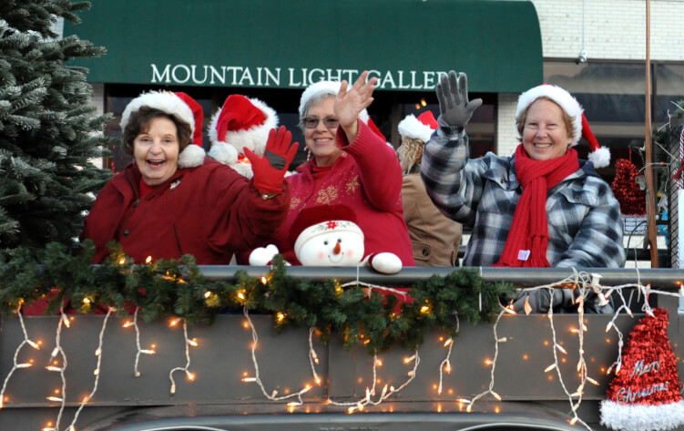 Three people in Christmas attire, waving from a decorated float with string lights and a snowman, in front of Mountain Light Gallery in Bishop, California. visit bishop