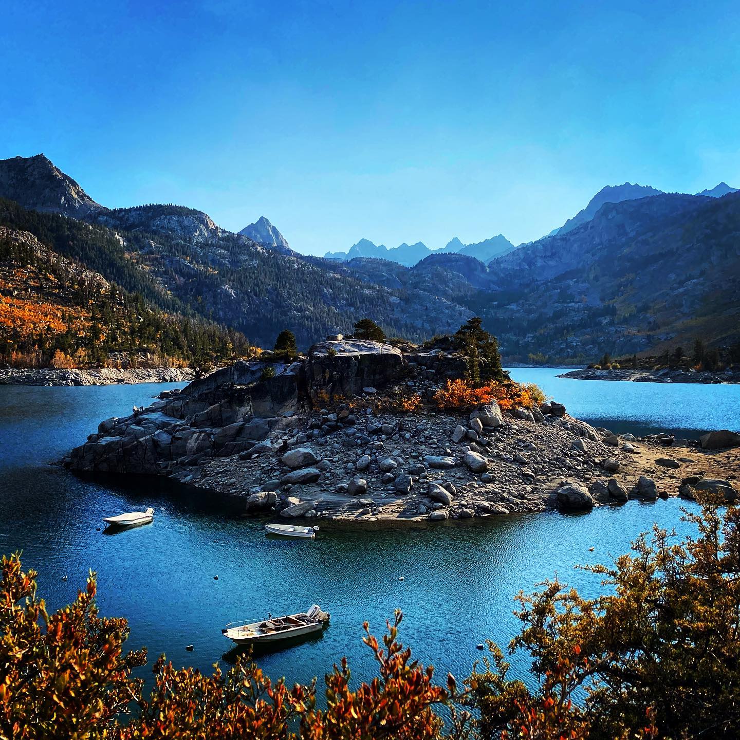 A serene lake near Bishop, California, surrounded by the majestic Eastern Sierra mountains, with a rocky island and three small boats floating in the calm blue water. visit bishop